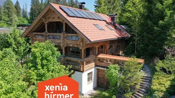 Traditionelles Holzhaus im alpinen Stil mit Ziegeldach und Solarpaneelen, umgeben von üppiger Natur.