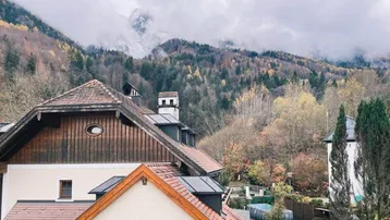 Blick von einem Balkon auf traditionelle Häuser, herbstliche Bäume und eine beeindruckende Berglandschaft.