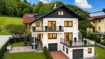 Modernes Einfamilienhaus mit weißer Fassade, großzügiger Terrasse, Balkonen und Doppelgarage, umgeben von grüner Natur.