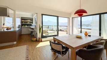 Heller offener Wohn- und Essbereich mit moderner Küche und direktem Zugang zur Terrasse mit See- und Bergblick.
