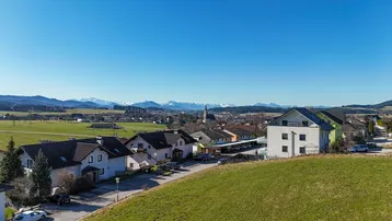 Panoramablick auf eine ländliche Wohnsiedlung mit grünen Feldern und schneebedeckten Bergen im Hintergrund.