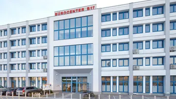 Modernes Bürogebäude mit weißer Fassade, blauen Fenstern und Parkplätzen davor.