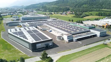 Luftaufnahme eines Gewerbeparks mit mehreren Gebäuden, die mit Photovoltaikanlagen auf den Dächern ausgestattet sind, umgeben von grünen Hügeln.