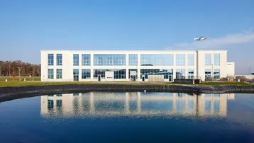 Modernes Geschäftsgebäude mit großen Glasflächen, reflektiert in einem Teich unter blauem Himmel mit Flugzeug.