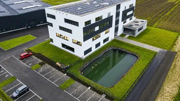 Modernes Geschäftsgebäude mit Solaranlage, Teich und Parkplätzen, umgeben von Grünflächen und weiteren Gewerbebauten.