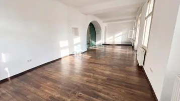 Geräumiger, heller Raum mit dunklem Holzfußboden, weißen Wänden und mehreren Fenstern, die viel Tageslicht hereinlassen.