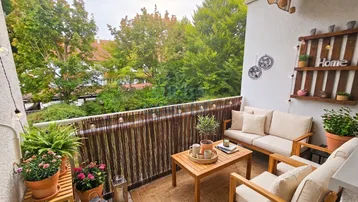 Möblierter Balkon mit gemütlichen Sitzgelegenheiten, vielen Pflanzen und Blick auf grüne Bäume.