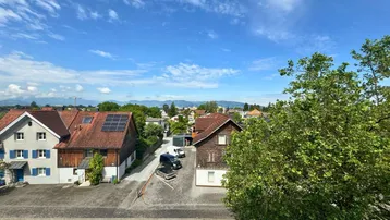 Weitblick über eine Wohnsiedlung mit Häusern, Bäumen und Bergen im Hintergrund unter blauem Himmel.