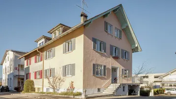 Traditionelles Wohnhaus mit hellrosa Fassade, grauen Fensterläden und Satteldach unter blauem Himmel.