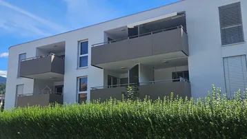 Modernes Mehrfamilienhaus mit mehreren Balkonen und gepflegter Hecke im Vordergrund.
