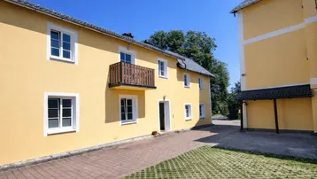 Gelbe Fassade eines Mehrfamilienhauses mit Balkon und gepflasterter Zufahrt unter blauem Himmel.