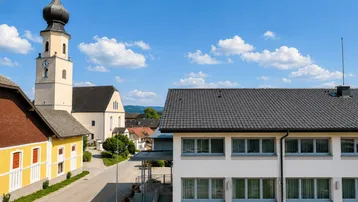 Blick auf ein österreichisches Dorf mit einer Kirche und einem modernen Wohngebäude unter blauem Himmel.