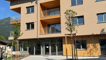 Modernes Mehrfamilienhaus mit beigefarbener Fassade, Holzverkleidung und Balkonen, umgeben von grüner Landschaft.
