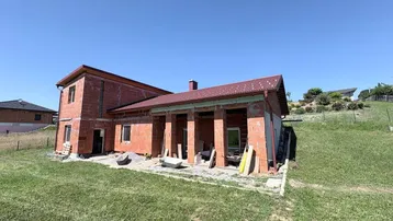 Haus im Rohbauzustand mit Ziegelwänden und rotem Dach, umgeben von einer grünen Wiese.