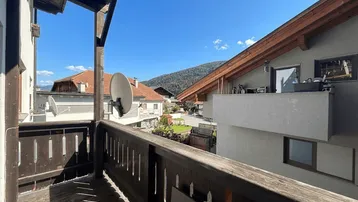 Traditioneller Holzbalkon mit Blick auf die umliegende Berglandschaft und benachbarte Gebäude unter blauem Himmel.
