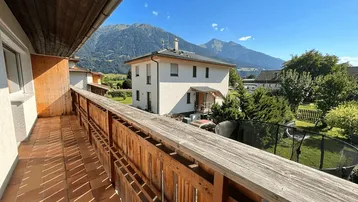 Heller Balkon mit Holzbalken, Fliesenboden und weitem Bergblick unter blauem Himmel.