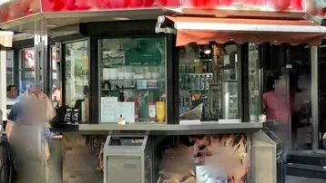 Ein kleiner, achteckiger Verkaufsstand mit rotem Dach und Glasfronten, verziert mit Flammenmotiven, in einer städtischen Umgebung.