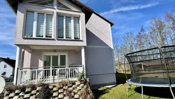 Hausfassade mit Balkon, Garten und Trampolin unter blauem Himmel an einem sonnigen Tag.