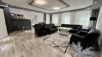 Geräumiges Wohnzimmer mit dunklen Sofas, hellem Laminatboden und moderner Deckenbeleuchtung.