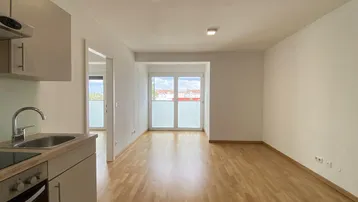 Heller offener Wohnbereich mit Holzboden, integrierter Küchenzeile und Zugang zu einem Balkon.
