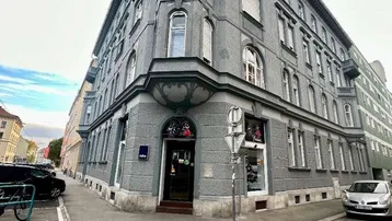 Graues Eckgebäude mit vielen Fenstern und Geschäften im Erdgeschoss an einer städtischen Straße.