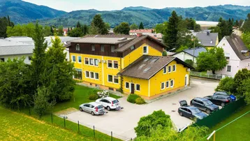 Gelbes Gebäudeensemble mit Parkplätzen und grüner Umgebung vor einer malerischen Bergkulisse.