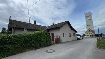 Älteres Haus mit grüner Hecke an einer Straße, im Hintergrund ein hoher Turm unter bewölktem Himmel.