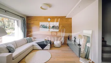 Helles Wohnzimmer mit Holzwand, großem Ecksofa und Blick ins Grüne durch die großen Fenster.