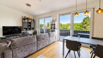 Heller Wohn- und Essbereich mit großem Sofa, Holzboden und Zugang zum Balkon mit grüner Aussicht.