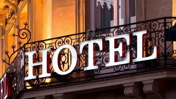 Abendliche Ansicht einer eleganten Hotelfassade mit leuchtendem Schriftzug und kunstvollem Balkongeländer.