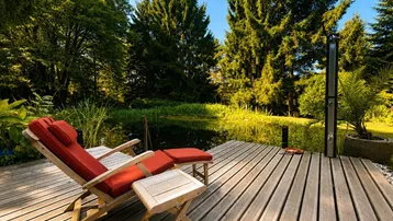 Holzterrasse mit Liegestuhl und Außendusche am Teich, umgeben von üppiger Natur und hohen Bäumen.
