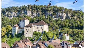 Malerische Burg über der Stadt, umgeben von bewaldeten Hügeln unter blauem Himmel.