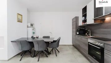 Moderne Küche mit grauen Schränken, integriertem Backofen und Essbereich mit dunklem Tisch.
