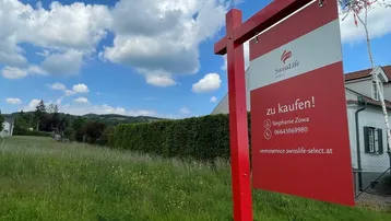 Rotes Verkaufsschild auf einem grünen Baugrundstück unter blauem Himmel mit einem Haus im Hintergrund.