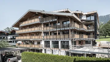 Großes Wohngebäude in Holzbauweise mit mehreren Balkonen, noch im Bau befindlich, vor alpiner Kulisse.