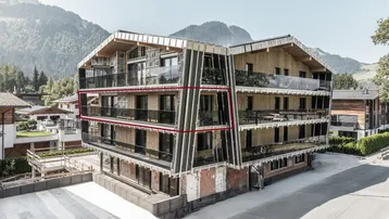 Modernes Mehrfamilienhaus in den Bergen mit Holzfassade und Glasbalkonen, umgeben von grüner Landschaft.