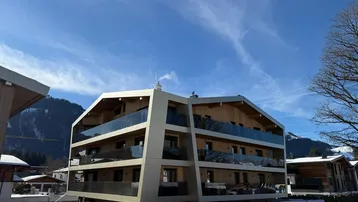 Modernes Mehrfamilienhaus in den Bergen mit Holzfassade und Glasbalkonen unter blauem Himmel.