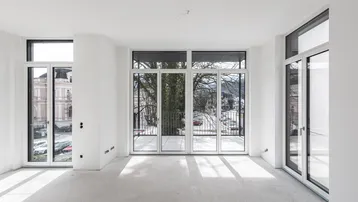 Heller, unmöblierter Raum mit großen Fenstern, hohen Decken und Zugang zu einem Balkon.