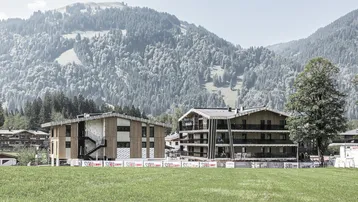 Zwei moderne Gebäude im Bau mit Holzfassaden und Bergblick in grüner Landschaft.
