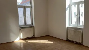 Helles, leeres Zimmer mit Parkettboden und großen Fenstern, das viel Tageslicht hereinlässt.