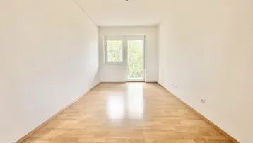 Helles, leeres Zimmer mit Parkettboden und Zugang zu einem Balkon.