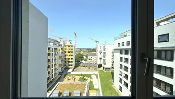 Blick aus dem Fenster auf moderne Wohngebäude, einen begrünten Innenhof mit Spielplatz und Baukräne.