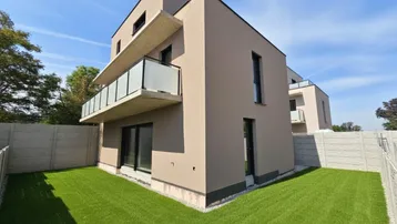 Modernes Gebäude mit beigefarbener Fassade, Balkon und kleinem Rasenbereich unter blauem Himmel.