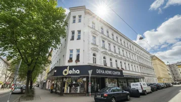 Historisches Eckgebäude mit Geschäften im Erdgeschoss und Wohnungen darüber unter strahlendem Sonnenschein.