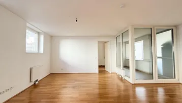 Heller Wohnbereich mit Parkettboden, weißen Wänden und Zugang zu einem verglasten Balkon.