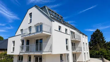Modernes Mehrfamilienhaus mit weißer Fassade, mehreren Balkonen und Dachfenstern unter blauem Himmel.