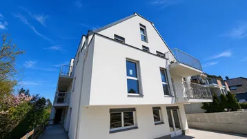 Moderne weiße Wohnanlage mit klaren Linien, großen Fenstern und Balkonen unter blauem Himmel.