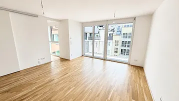 Helles, leeres Wohnzimmer mit Holzboden und Zugang zu einem Balkon in einem Neubau.
