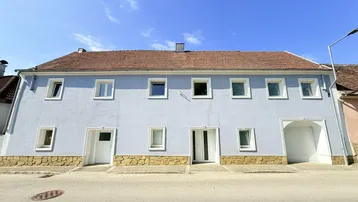 Hellblau gestrichene Fassade eines Mehrfamilienhauses mit braunem Ziegeldach und Steinsockel.