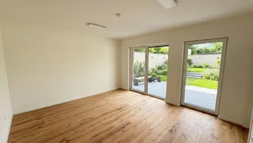 Helles, leeres Zimmer mit Holzboden und großen Fenstern zum gepflegten Gartenbereich.